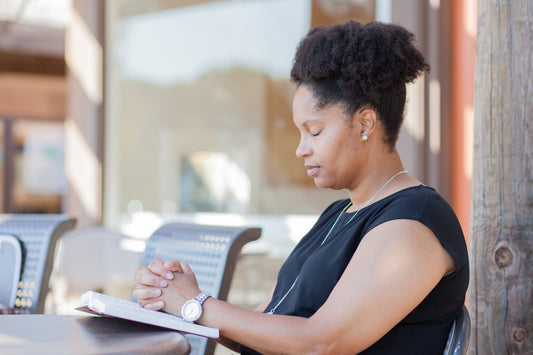A Black woman sits outdoors with her eyes closed and hands clasped in prayer over an open book.