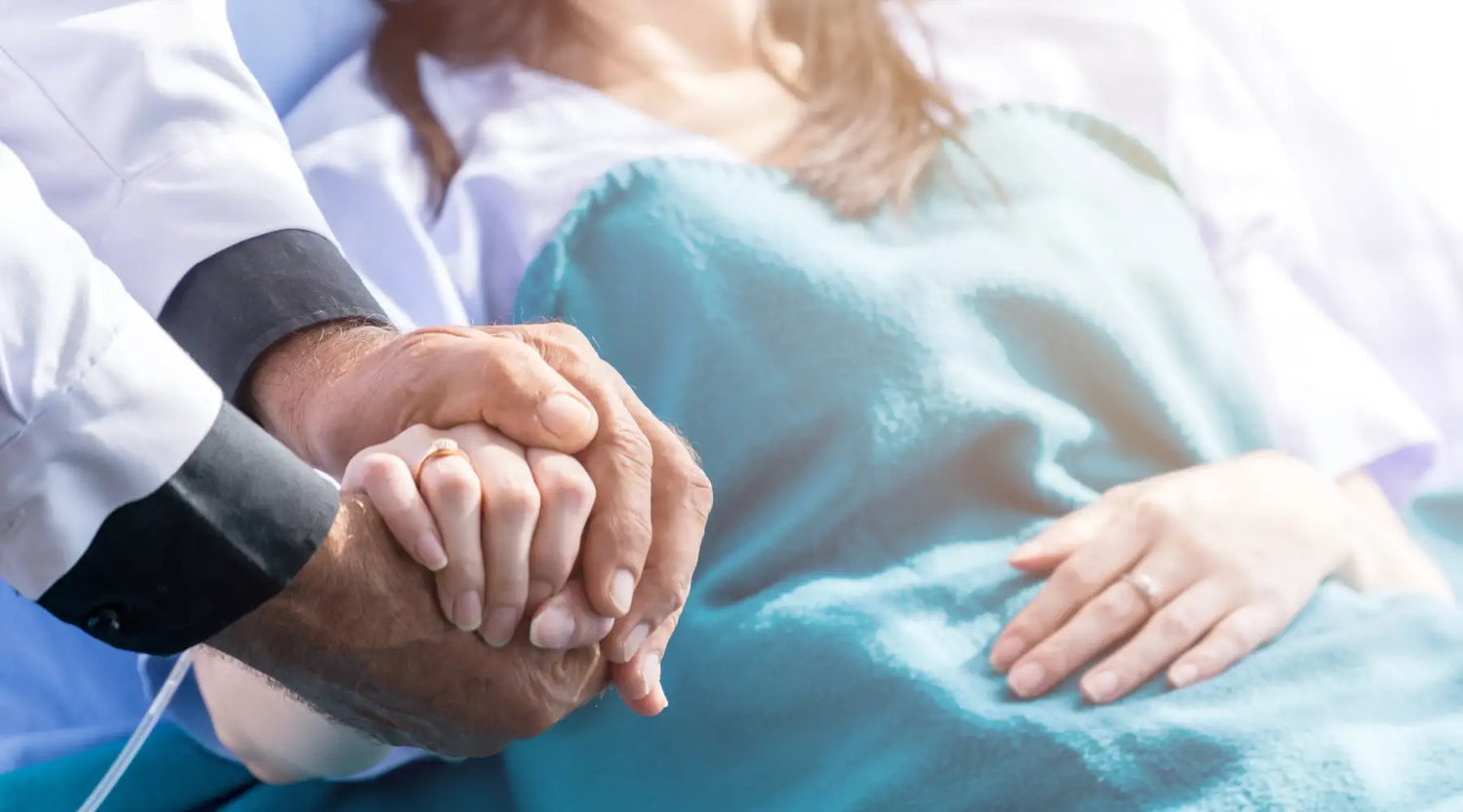 A doctor’s hand, wearing a white coat with black cuffs, gently holds the patient’s hand.