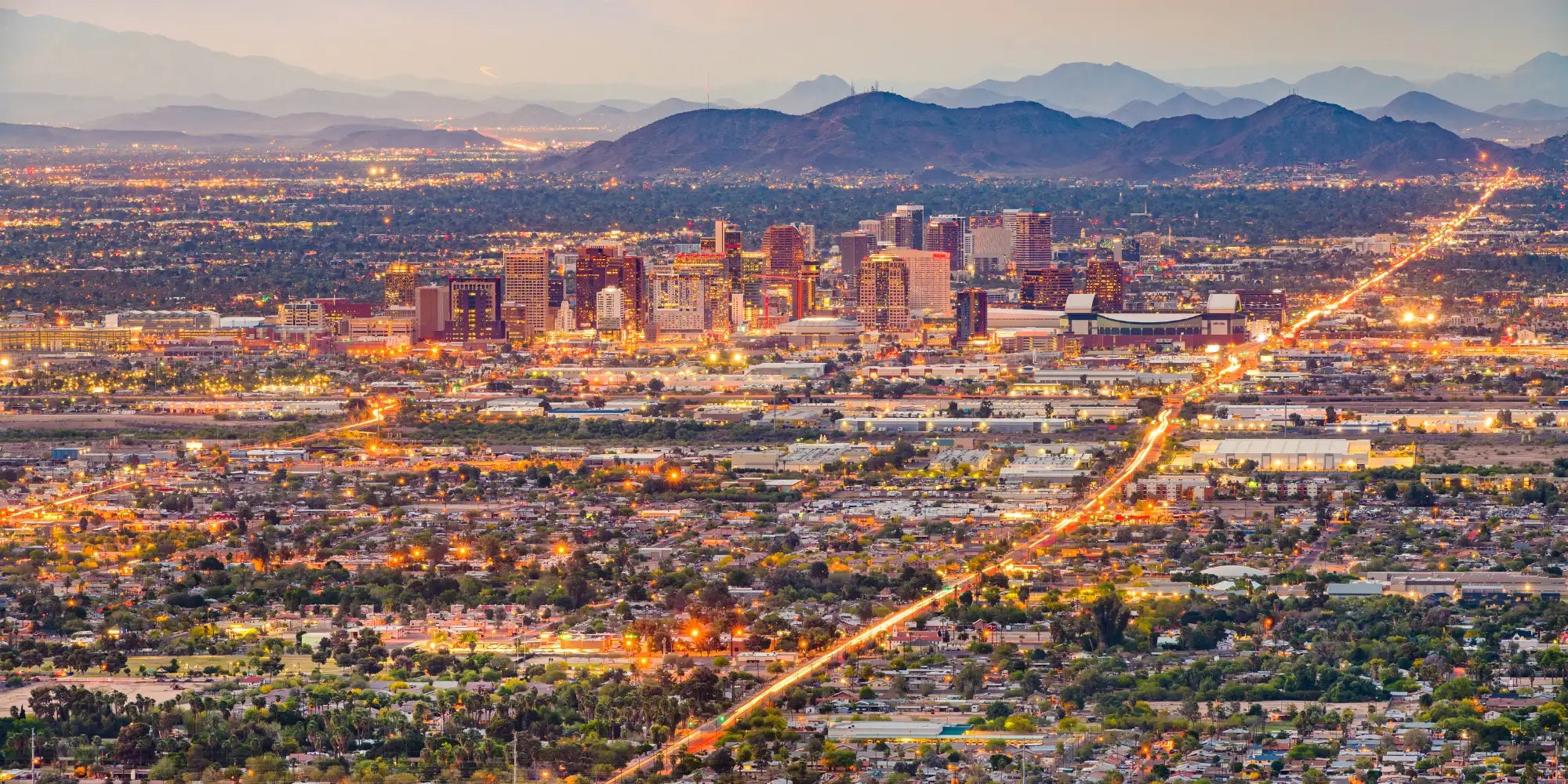 The illuminated skyline of phoenix at dusk, featuring modern high-rise buildings with warm golden and orange lights against a backdrop of distant mountains.