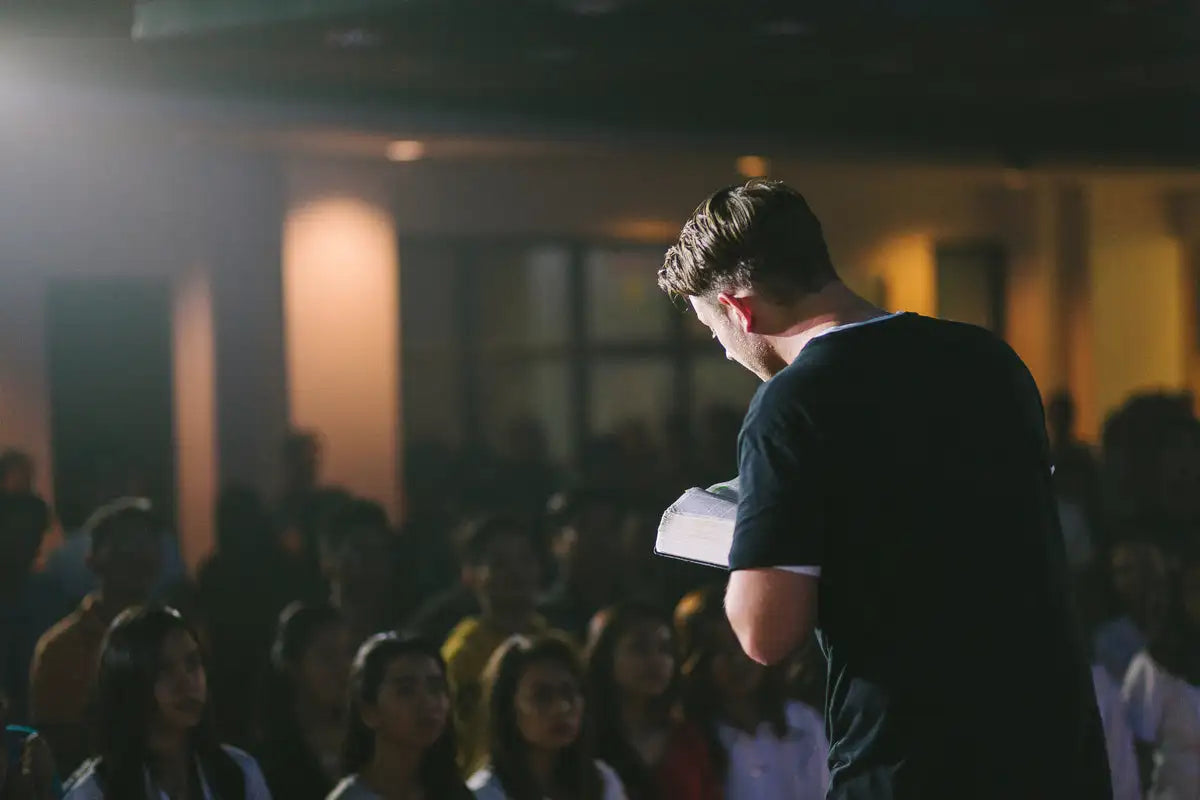 A man in a black t-shirt stands before an audience, holding a white book.