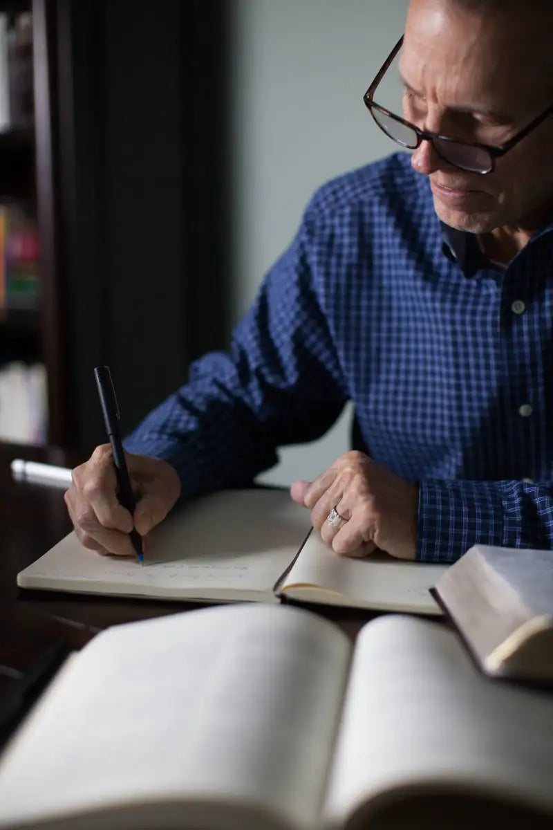 Man wearing a blue plaid button-up shirt and glasses, writing in an open notebook with a black pen.