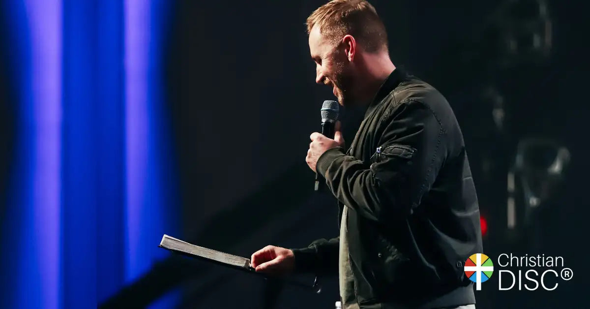 A man in a dark jacket holding a black microphone and an open booklet.