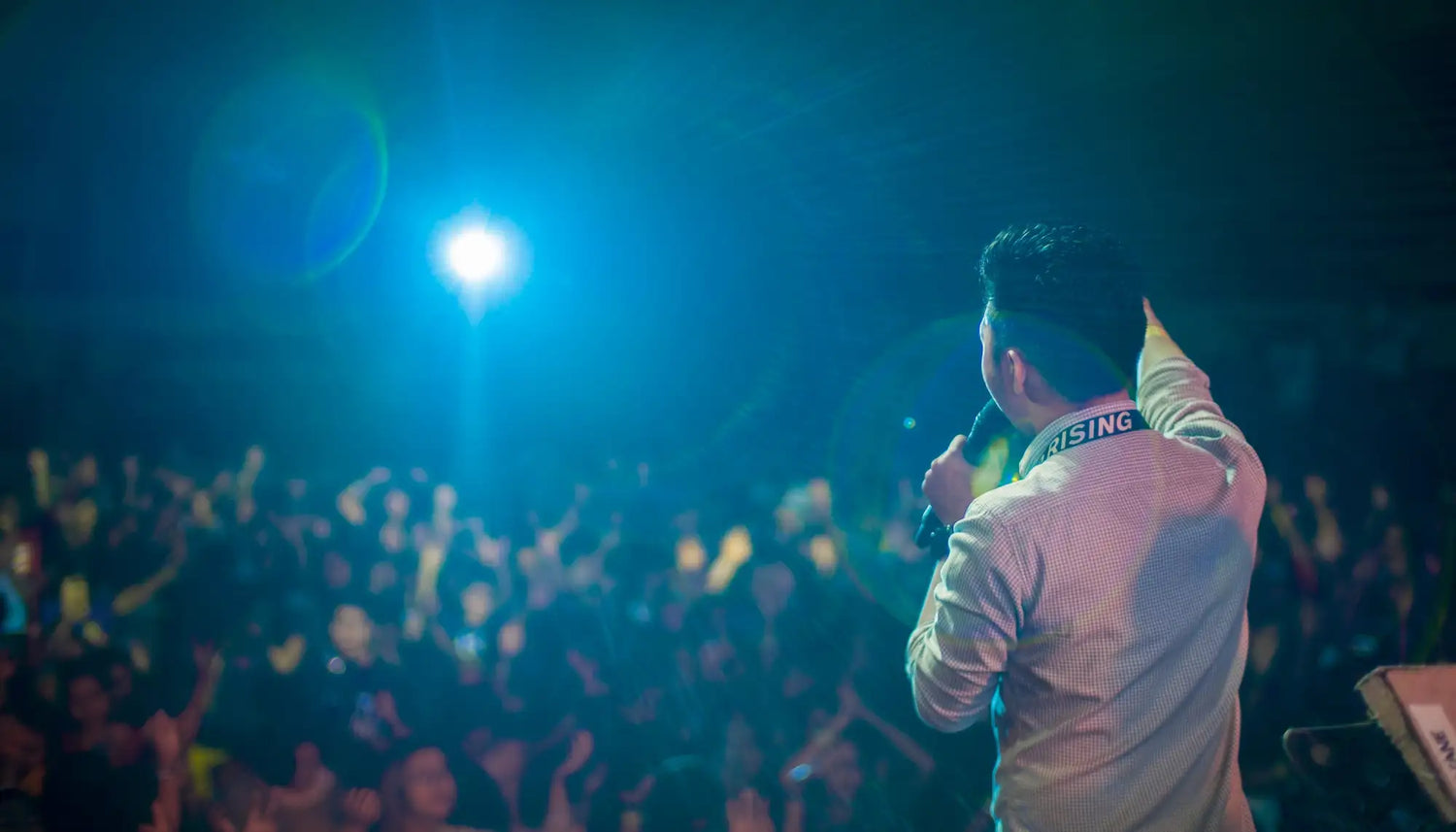 A man in a light-colored, subtly patterned shirt stands on stage holding a microphone with his left hand raised.