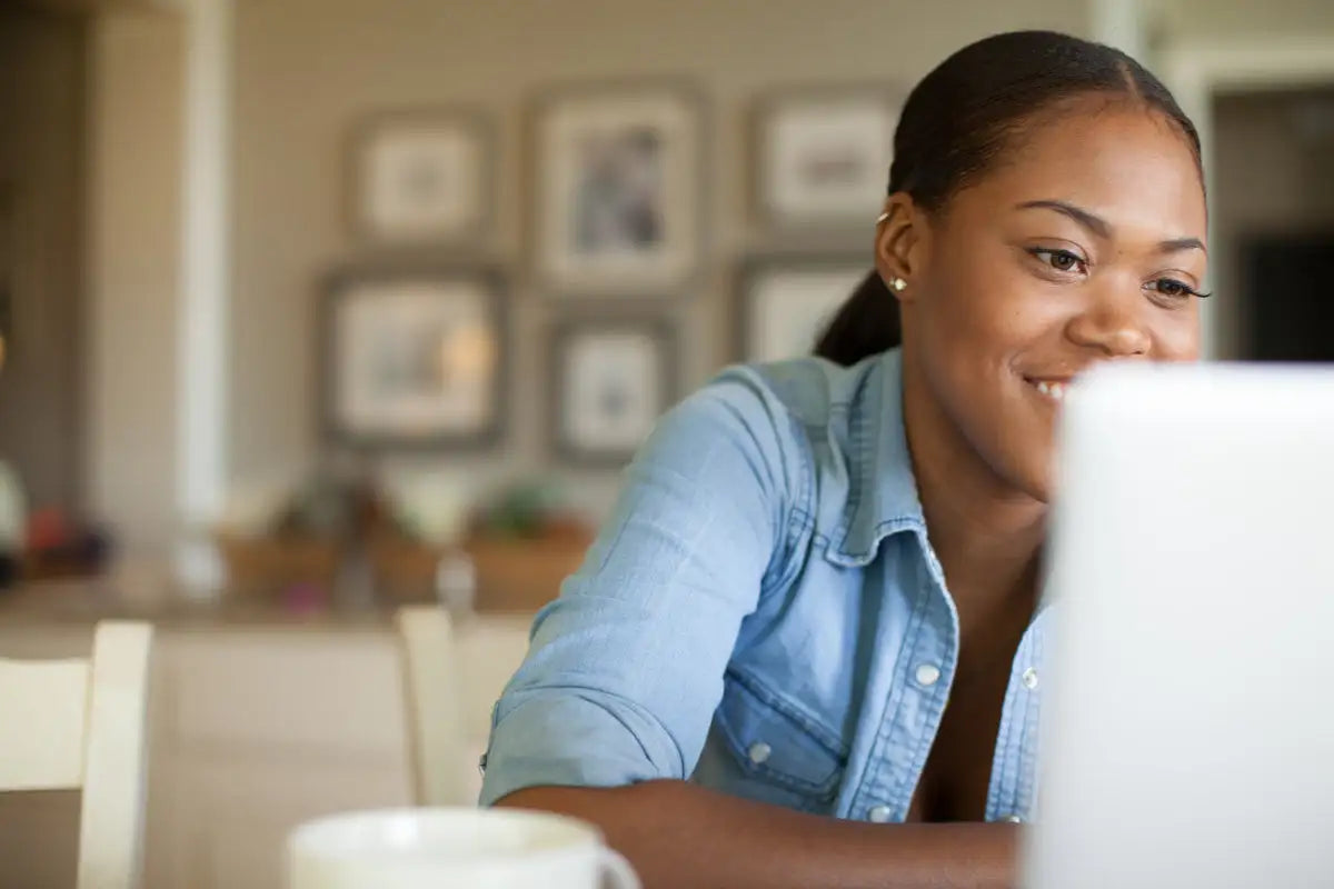 A woman wearing a light blue denim shirt sits at a table with a white laptop in front of her.